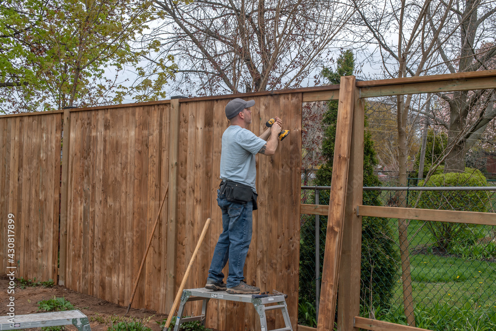 A white, middle-aged gay man builds a wooden fence in his back yard.