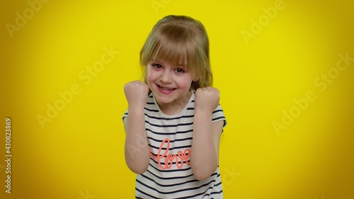 Portrait of funny playful blonde kid child shouting, raising hands in gesture I did it, celebrating success, winning and goal achievemen on yellow studio background. Teenager children girl emotions