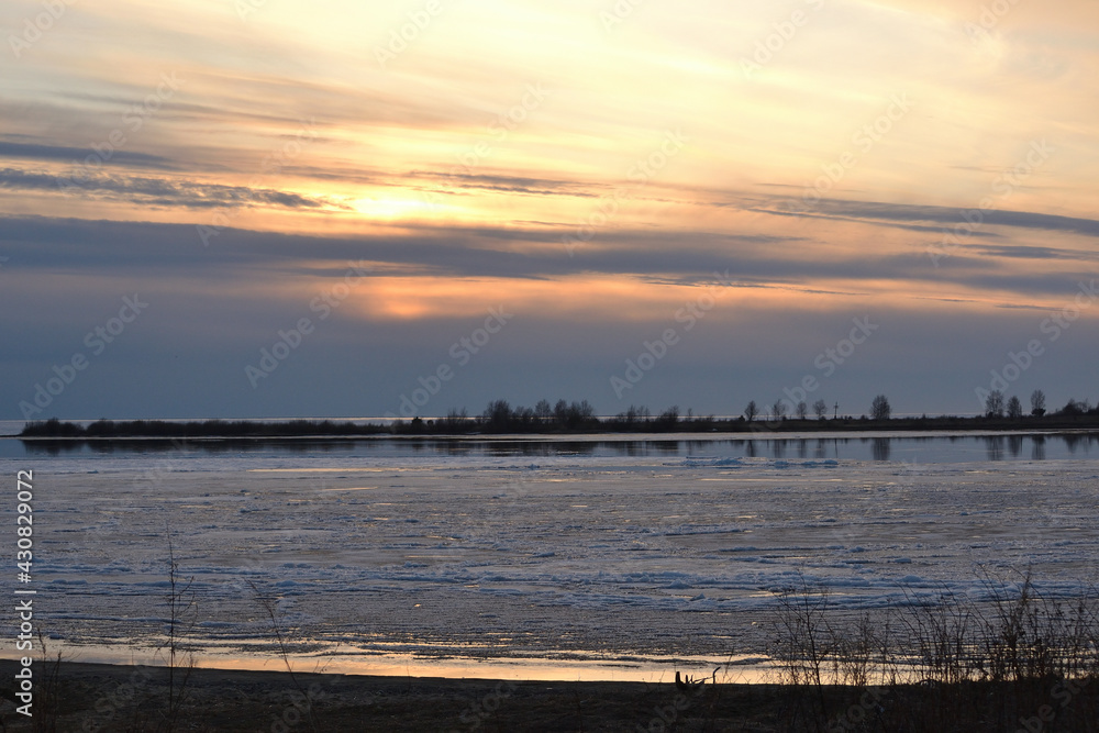 A landscape view of the frozen sea during ice drift in spring with dramatic sky and with reflections in melt water in the evening at sunset. In the distance, the black strip of the island.