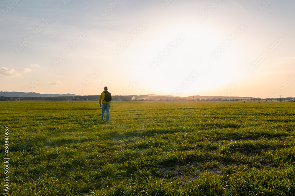 Young caucasian man in yellow jacket with backpack standing on meadow, field at sunset in czech spring landscape