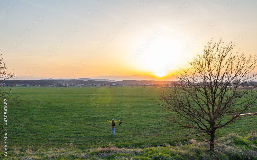 Fototapeta premium Aeirial view young caucasian man in yellow jacket with backpack standing on meadow, field at sunset in czech spring landscape