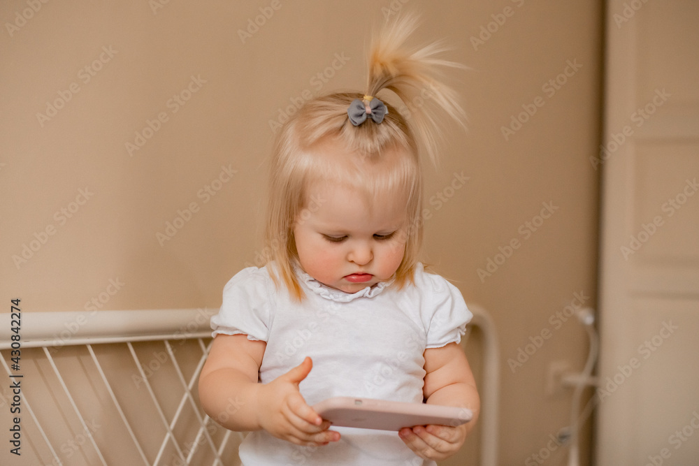 Fototapeta premium Baby blonde girl plays with the phone at home on the bed. Children and technology