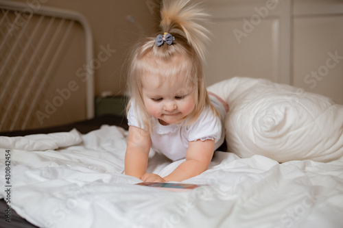 Baby blonde girl plays with the phone at home on the bed. Children and technology