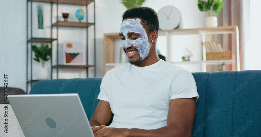 Handsome positive young modern black-skinned guy with moisturiser face mask sitting in front of laptop on the sofa and chatting with friends Handsome positive young modern black-skinned guy with moisturiser face mask sitting in front of laptop on the sofa and chatting with friends