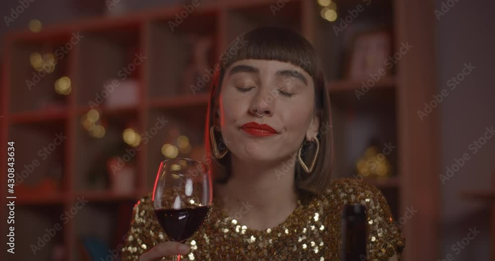 Joyful woman drinking from wineglass and smiling while having online conversation. Cheerful female person laughing while looking to camera and sitting at home. Concept of online date. Joyful woman drinking from wineglass and smiling while having online conversation. Cheerful female person laughing while looking to camera and sitting at home. Concept of online date.