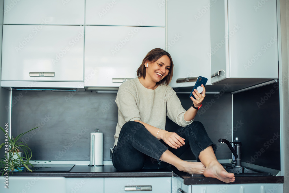 Cheerful brunette sits on countertop in a home kitchen with smartphone in her hand and laughs. Communication with friends and family in conditions of self-isolation and quarantine. Cheerful brunette sits on countertop in a home kitchen with smartphone in her hand and laughs. Communication with friends and family in conditions of self-isolation and quarantine.