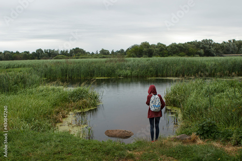 Slim girl teenager tourist in a hood stands on the shore of a lake of a river in a reservoir in summer. Rainy cool cloudy weather.