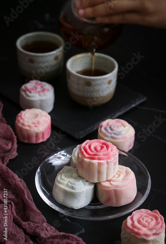 A plate of colorful pastel traditional non-baked Asian snow skin mooncake with cup and hand pouring tea in background. Usually eat chilled and served to celebrate Mid-Autumn festival 