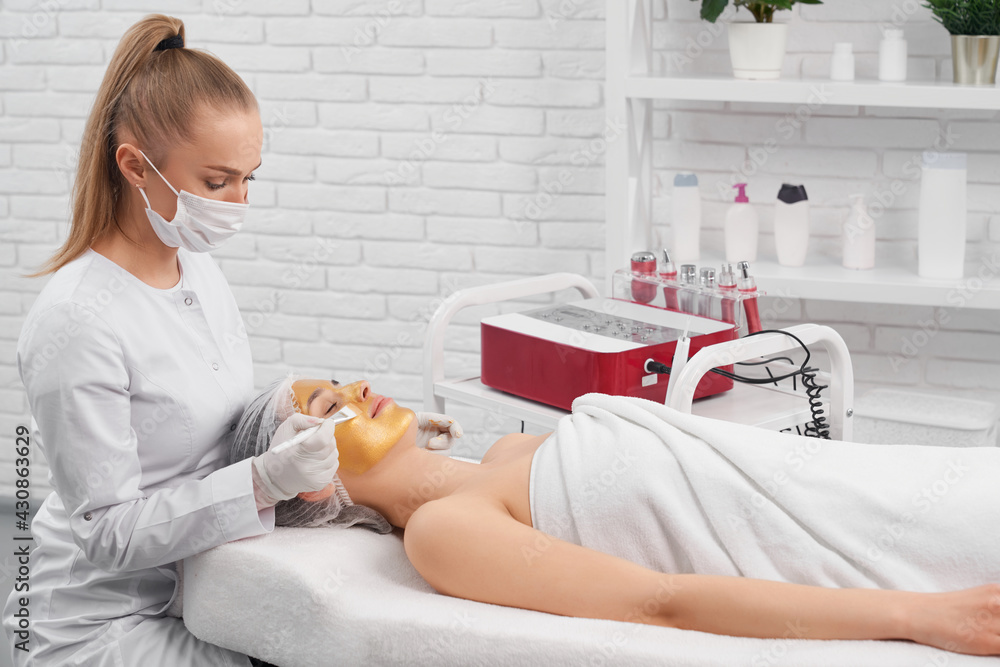 Side view of young beautician in protective mask and rubber gloves doing special cosmetic mask for young beautiful patient. Concept of procedure for improvements skin in professional salon. Side view of young beautician in protective mask and rubber gloves doing special cosmetic mask for young beautiful patient. Concept of procedure for improvements skin in professional salon.
