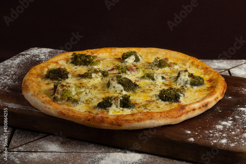 One whole broccoli pizza on wooden board and table with bacon and wheat around