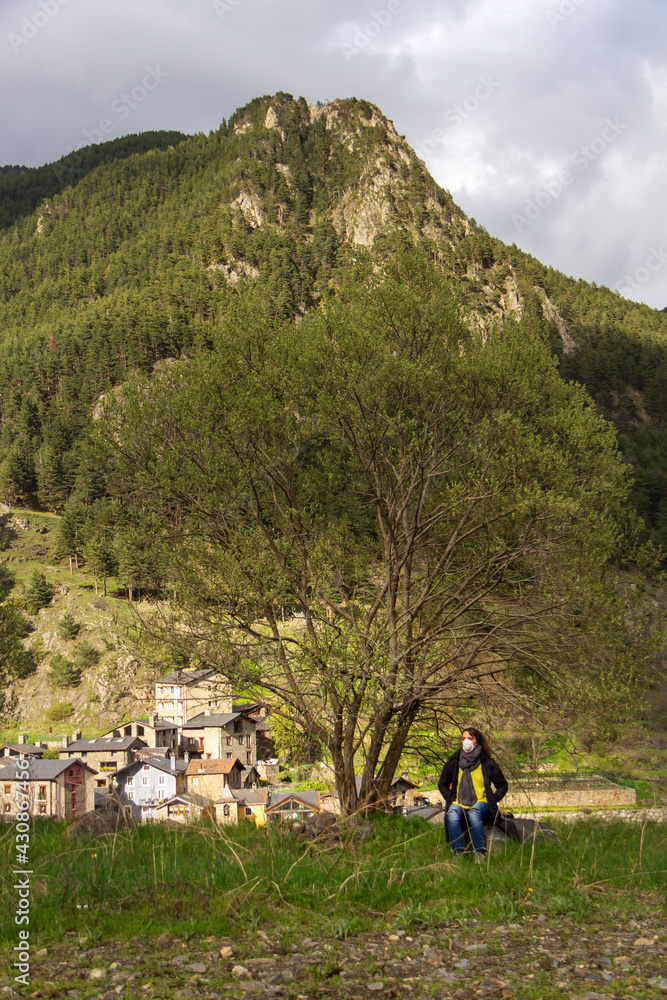 woman with mask and black coat rests on a stone under a tree in the countryside with the mountain in the background woman with mask and black coat rests on a stone under a tree in the countryside with the mountain in the background