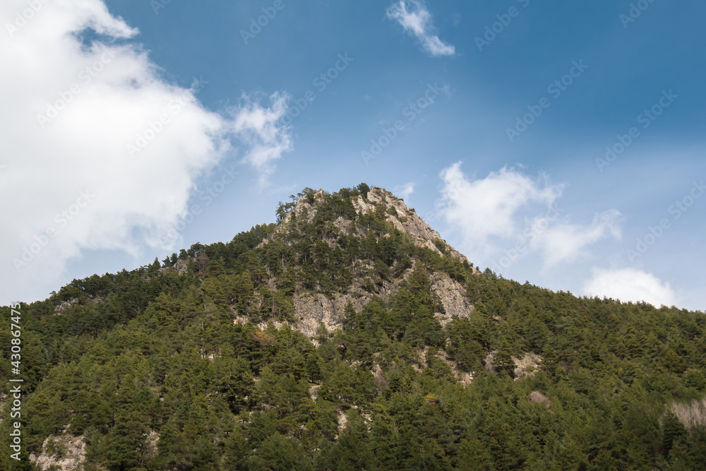 a beautiful mountain surrounded by green trees and under a blue sky with a big cloud on the side.