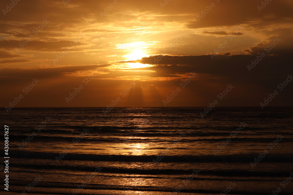 Fototapeta premium Sunset on a beach, orange sun is shining through the clouds and reflected in dark waves. Evening sea background for romantic travel