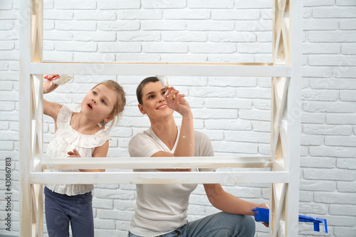 Close up of mother with little daughter painting wooden rack in white color. Concept of repair at home.