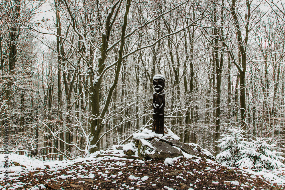 Carved wooden statue covered with snow standing on small hill in winter forest. Wooden sculpture of old Celt.Vacation outdoors harmony with winter nature landscape. Faith motivation background.