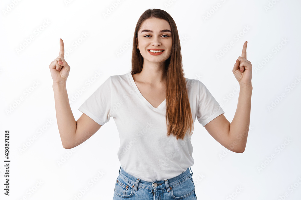 Smiling beautiful girl proudly showing advertisement, pointing fingers up at top promo deal, show discounts shop, standing over white background in t-shirt Smiling beautiful girl proudly showing advertisement, pointing fingers up at top promo deal, show discounts shop, standing over white background in t-shirt