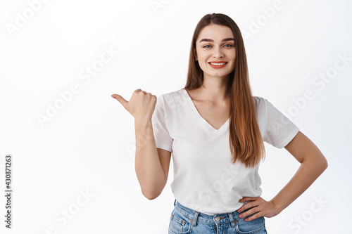 Smiling cheerful woman points thumb left, shows copy space advertisement, store promo deal, look determined, recommend product, stands against white background