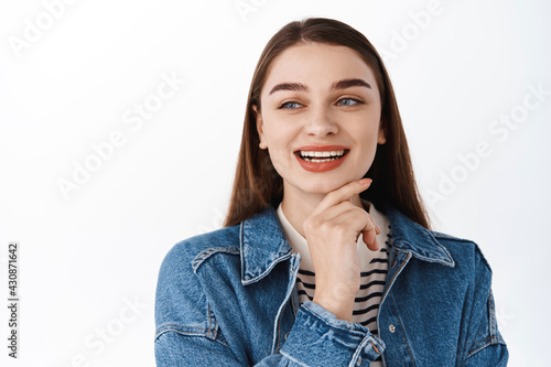 Thoughtful happy girl looking away at copy space with promotional text, smiling and touching chin while thinking, consider to buy product on left advertisement place, standing over white background