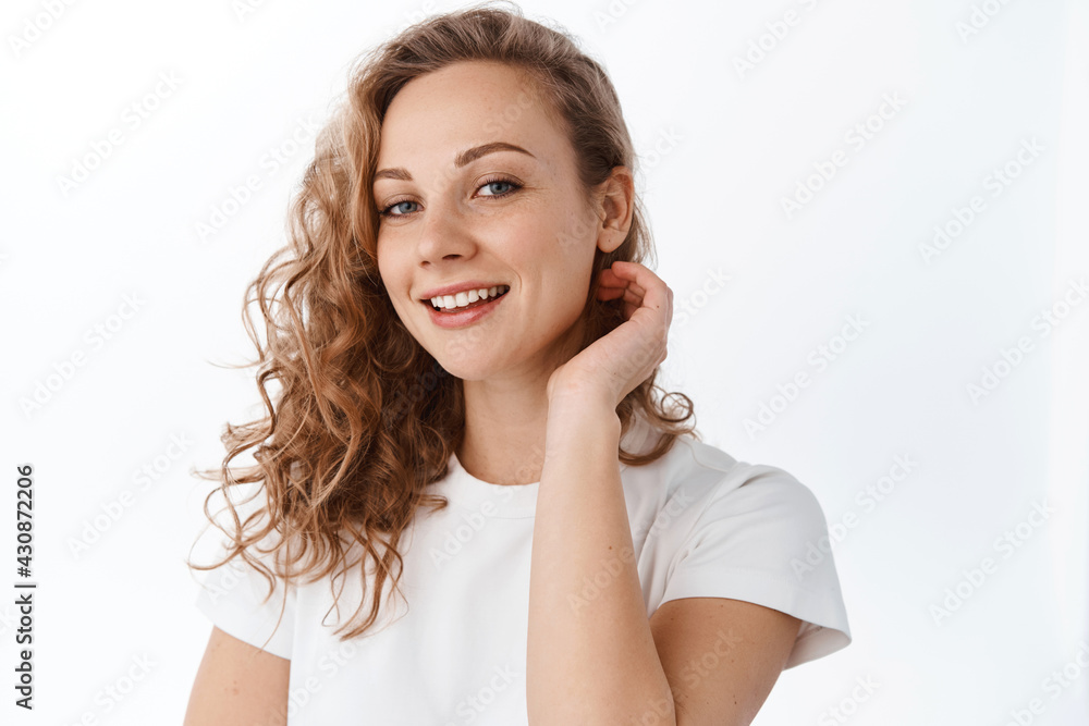 Young woman with curly blond hairdo, tuck hair behind ear and smile happy at camera, look natural and relaxed, standing over white background Young woman with curly blond hairdo, tuck hair behind ear and smile happy at camera, look natural and relaxed, standing over white background