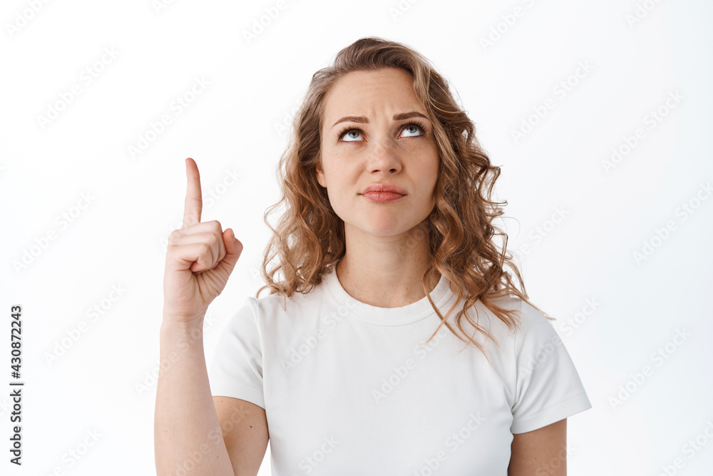 Skeptical and doubtful blond woman grimacing, pointing and looking up unsure, standing against white background Skeptical and doubtful blond woman grimacing, pointing and looking up unsure, standing against white background