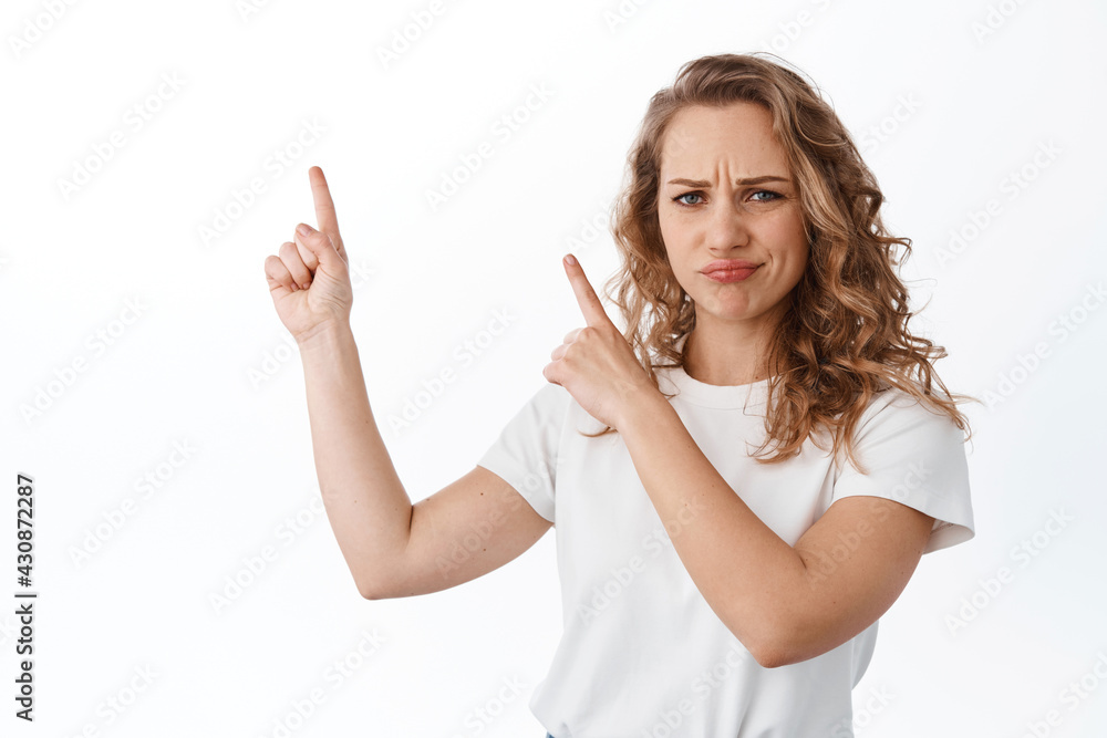 Skeptical blond girl complaining, frowning and sulking judgemental, pointing fingers at upper left corner logo, showing promo text, standing over white background Skeptical blond girl complaining, frowning and sulking judgemental, pointing fingers at upper left corner logo, showing promo text, standing over white background