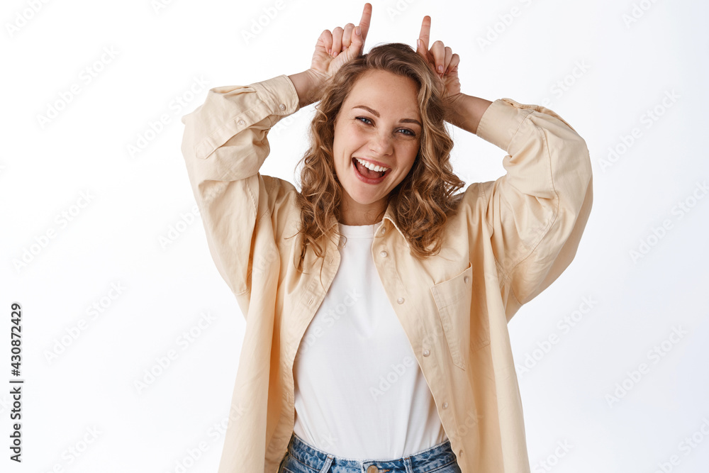 Naklejka premium Portrait of cheerful blond girl stay positive, showing stubborn horns gesture on head, smiling and looking happy, standing over white background