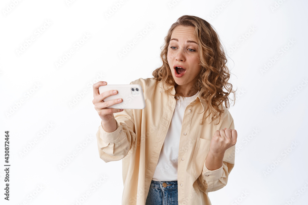 Girl looks at smartphone with amazement, watching video or stream, checking out awesome thing online, standing with mobile phone over white background Girl looks at smartphone with amazement, watching video or stream, checking out awesome thing online, standing with mobile phone over white background