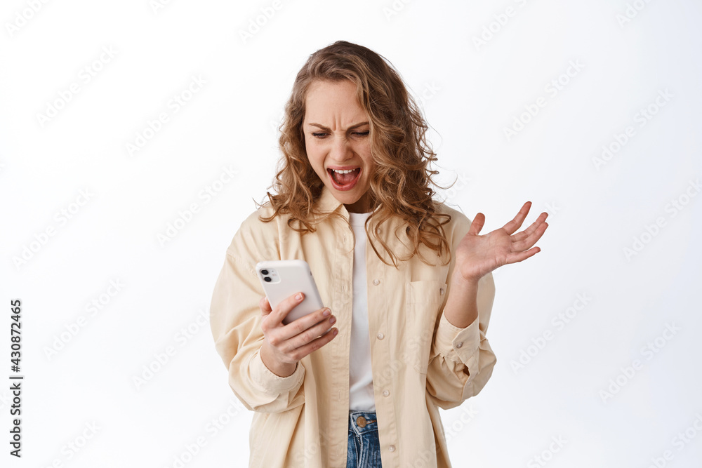 Angry woman looking at smartphone screen and complaining, looking furious at mobile phone, standing over white background Angry woman looking at smartphone screen and complaining, looking furious at mobile phone, standing over white background