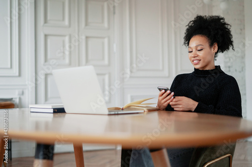 In the browser, working on a new work project. A satisfied female designer student smiles at an Afro-ethnic appearance. Prints a message on the phone in the messenger. There is a laptop on the table. © muse studio