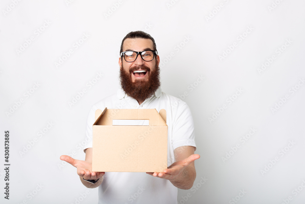 An excited young man with a delivery box is smiling amazingly at the camera near white wall An excited young man with a delivery box is smiling amazingly at the camera near white wall