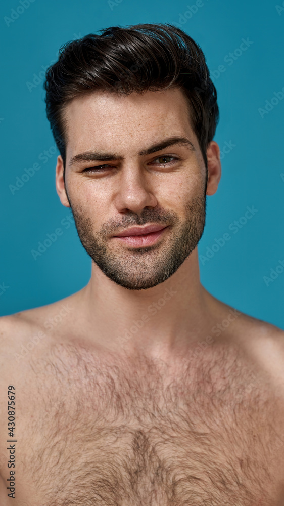 Portrait of attractive naked brunette man with fresh perfect smooth skin screw up one eye while looking at camera isolated over blue background Portrait of attractive naked brunette man with fresh perfect smooth skin screw up one eye while looking at camera isolated over blue background