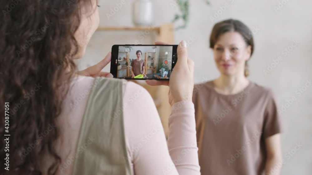Over-the-shoulder medium rear-view shot of woman with long curly hair holding smartphone, filming video of her female friend telling how to make natural organic cosmetics at home
