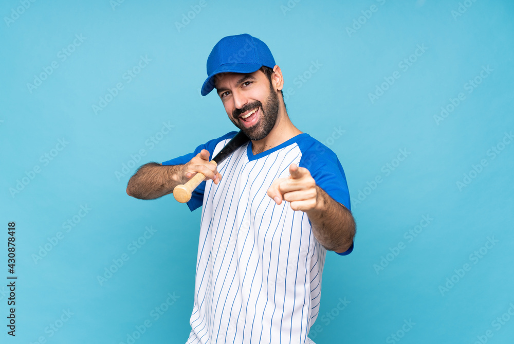 Young man playing baseball over isolated blue background points finger at you while smiling