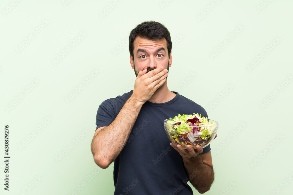 Young handsome man with salad over isolated green wall covering mouth with hands Young handsome man with salad over isolated green wall covering mouth with hands