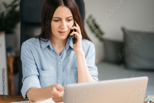 Attractive business woman sits at table in front of laptop and uses smartphone for work. Beautiful brunette girl is undergoing online training while at home. Female millennial surfing the internet