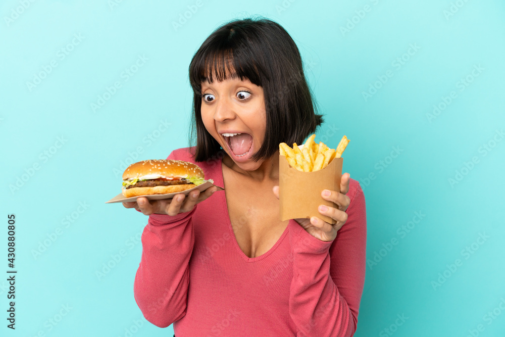 Young brunette woman holding burger and fried chips over isolated blue background