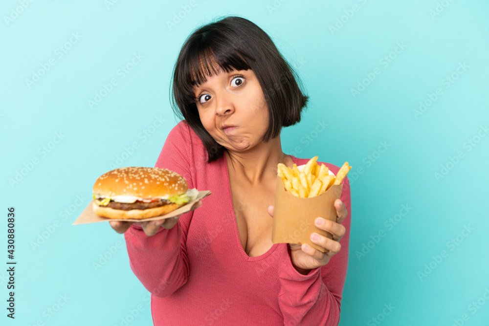 Young brunette woman holding burger and fried chips over isolated blue background Young brunette woman holding burger and fried chips over isolated blue background
