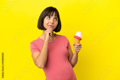 Pregnant woman holding a cornet ice cream isolated on yellow background and looking up © luismolinero
