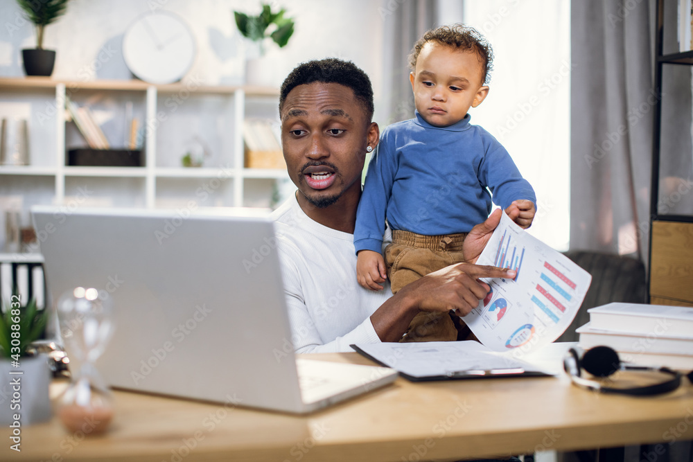 Afro american businessman having online meeting on modern laptop while carrying his baby son on hands. Young father working remotely from home. Afro american businessman having online meeting on modern laptop while carrying his baby son on hands. Young father working remotely from home.