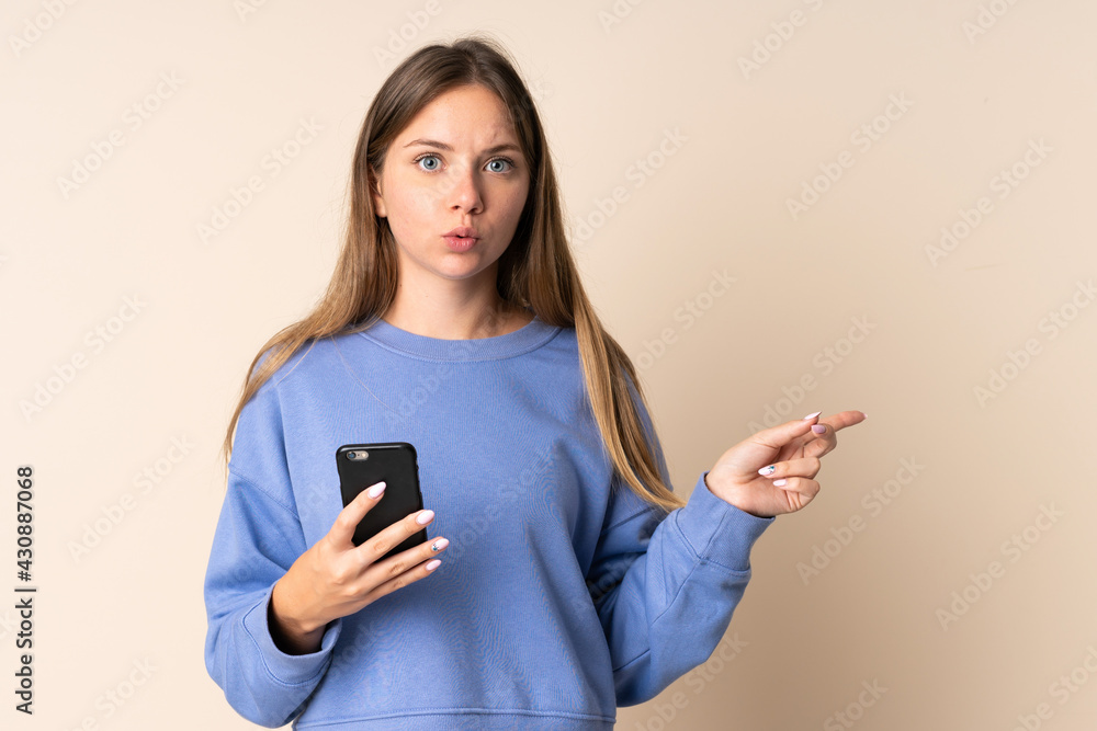 Young Lithuanian woman using mobile phone isolated on beige background surprised and pointing side
