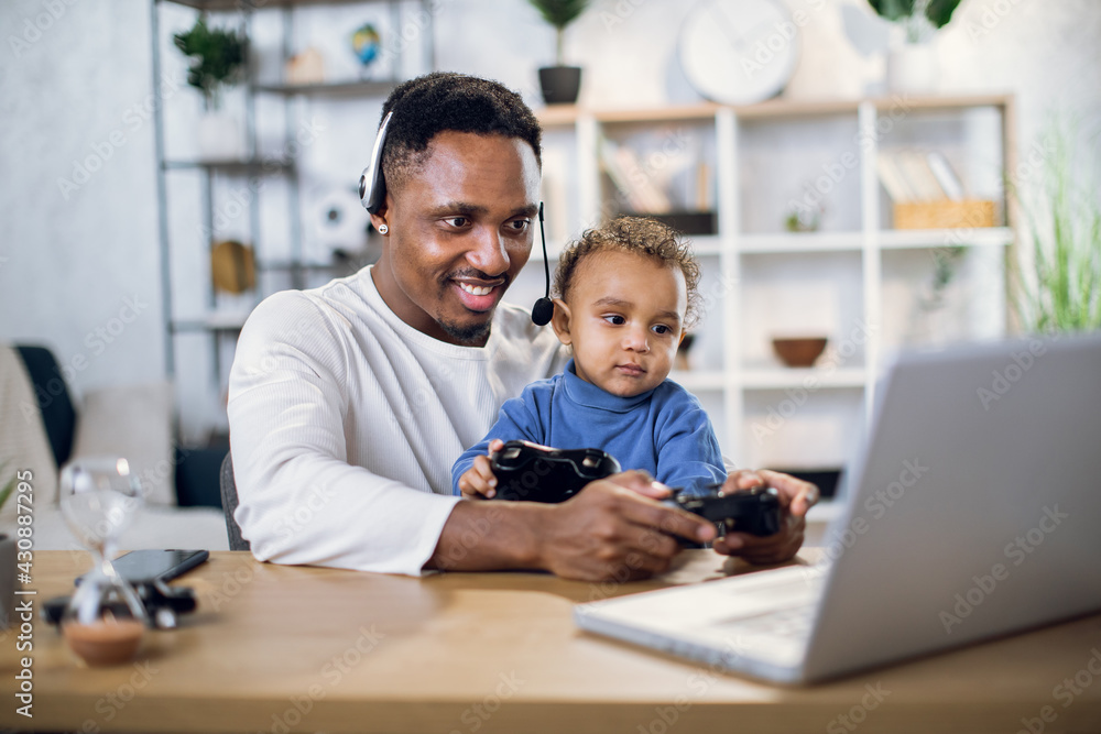 Afro american father and cute son using modern laptop and wireless joysticks for playing online games. Concept of entertainment, family and leisure time. Afro american father and cute son using modern laptop and wireless joysticks for playing online games. Concept of entertainment, family and leisure time.