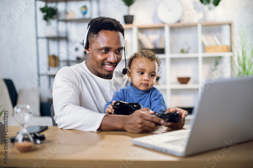 Afro american father and cute son using modern laptop and wireless joysticks for playing online games. Concept of entertainment, family and leisure time. © sofiko14