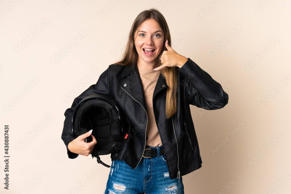 Young Lithuanian woman holding a motorcycle helmet isolated on beige background making phone gesture. Call me back sign Young Lithuanian woman holding a motorcycle helmet isolated on beige background making phone gesture. Call me back sign