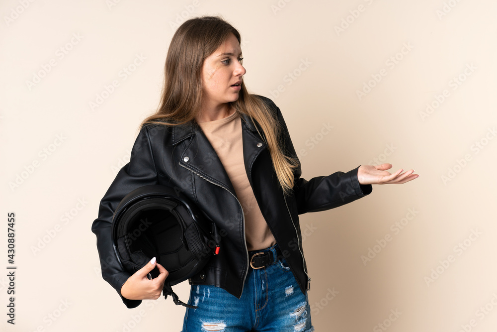 Young Lithuanian woman holding a motorcycle helmet isolated on beige background with surprise expression while looking side Young Lithuanian woman holding a motorcycle helmet isolated on beige background with surprise expression while looking side
