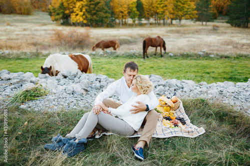 Couple is sitting hugging each other on a blanket with food on the lawn. Horses graze in the background