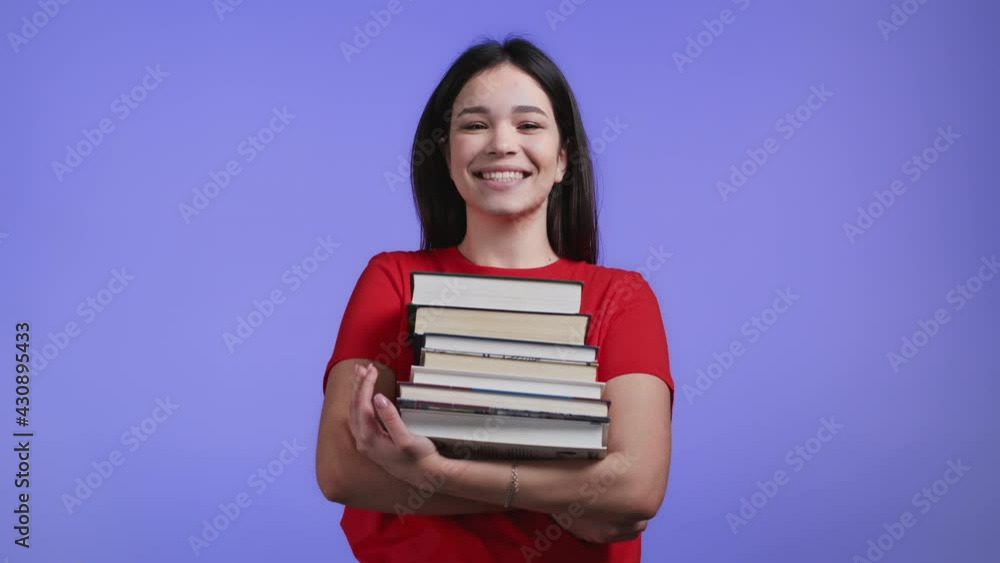 Student girl holds stack of university books from library on violet background in studio. Woman smiles, she is happy to graduate. Student girl holds stack of university books from library on violet background in studio. Woman smiles, she is happy to graduate.