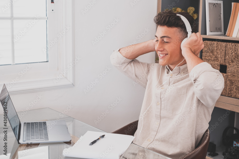 Happy relaxed millennial teenager business man wear wireless headphones look away rest at workplace finished work listening music podcast feel peace of mind concept sit at desk in office writing. Happy relaxed millennial teenager business man wear wireless headphones look away rest at workplace finished work listening music podcast feel peace of mind concept sit at desk in office writing.