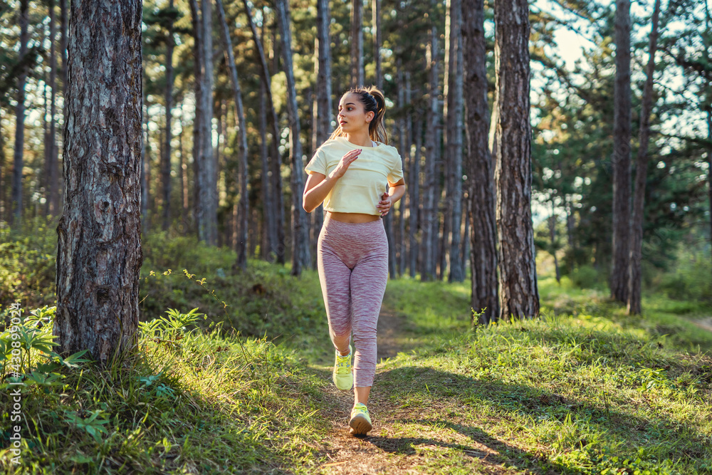 Front view full length of young caucasian woman running trough the woods in nature outdoor jogging in sunny day - sport fitness and recreation concept