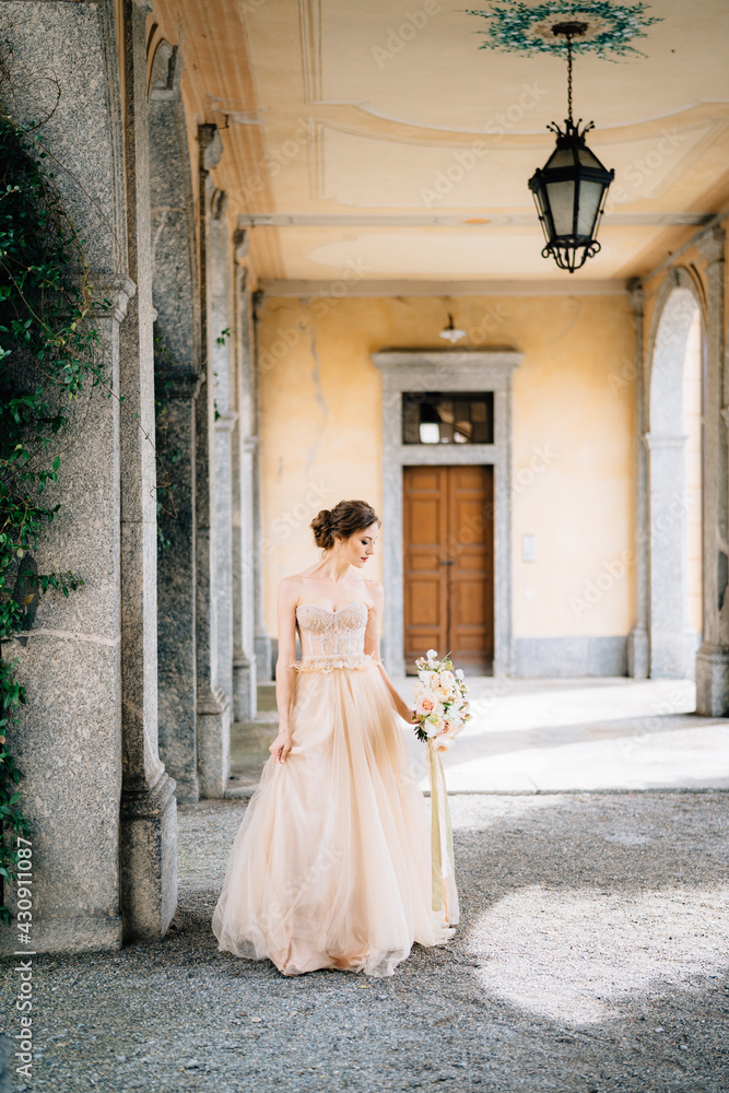 Bride in a beautiful dress with a bouquet of pink flowers stands in an arched corridor near the greenery. Lake Como, Italy