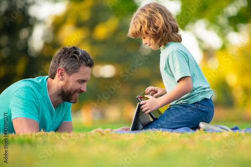 Happy father using laptop relax with schooler son holding laptop have fun together, smiling dad and little boy child enjoy weekend with gadgets outside on nature. Video call of a grandson to parents.